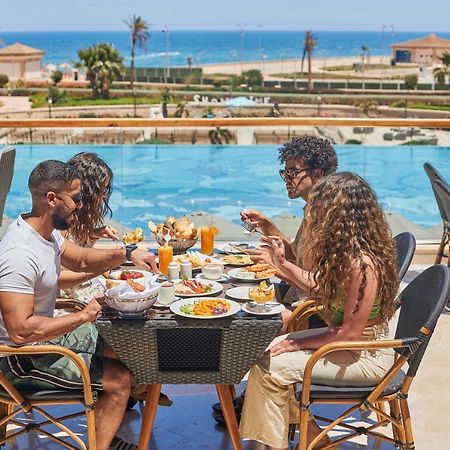 Tolip Resort El Galala Majestic Ain Sukhna Exterior foto The photo shows a group of five people enjoying a meal together outdoors, likely at a restaurant or cafe. They are seated at a table with a beautiful view of a swimming pool and the sea in the background. The table is filled with various dishes, and 