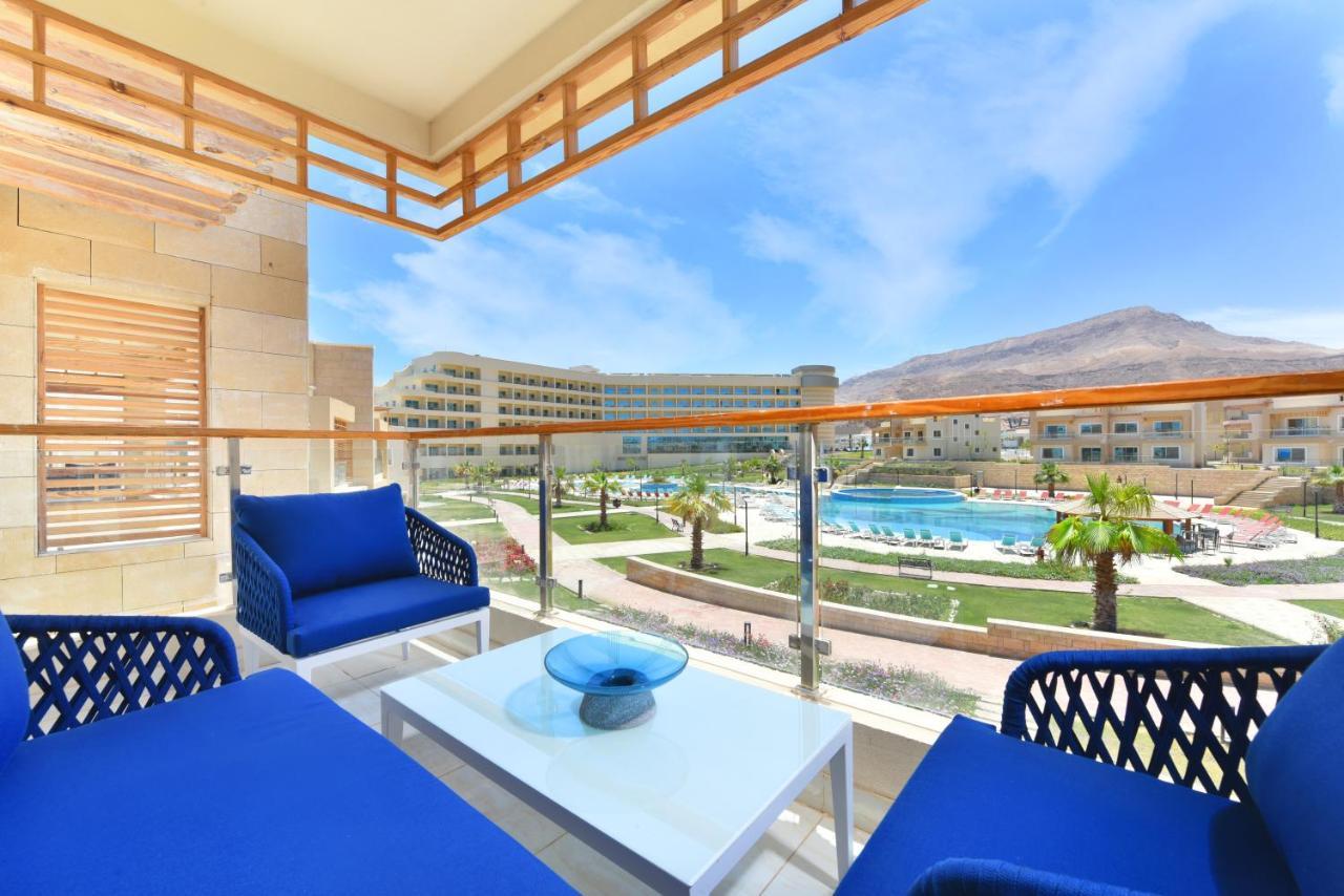 Tolip Resort El Galala Majestic Ain Sukhna Exterior foto The photo showcases a balcony view featuring two blue chairs and a table with a decorative bowl. The balcony overlooks a landscape that includes a swimming pool and manicured lawns. In the background, there are multiple buildings and palm trees, all 