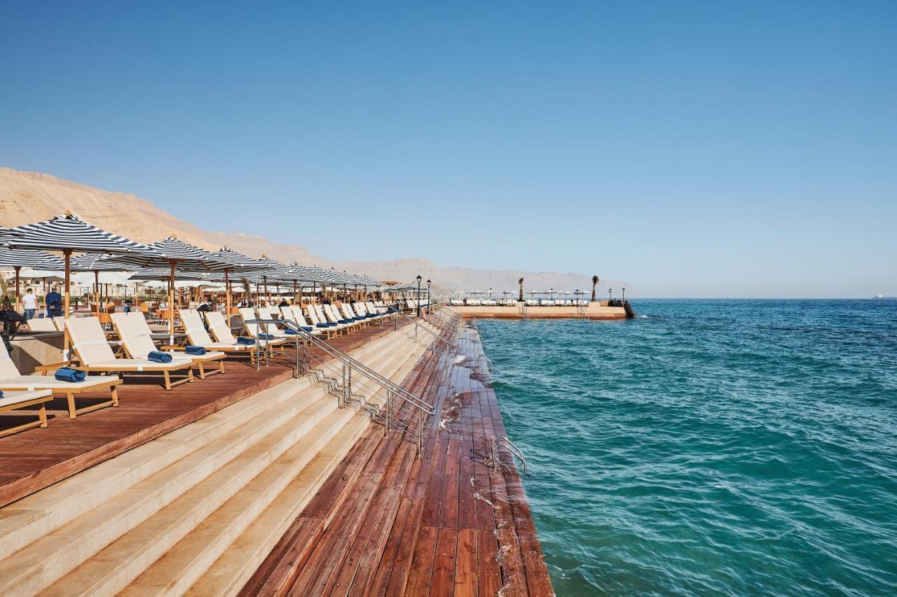 Tolip Resort El Galala Majestic Ain Sukhna Exterior foto The photo shows a seaside area featuring a wooden deck that extends into the water. On the left side, there are several lounge chairs arranged neatly under shade structures, suggesting a relaxing atmosphere. The ocean is visible in the foreground, wi