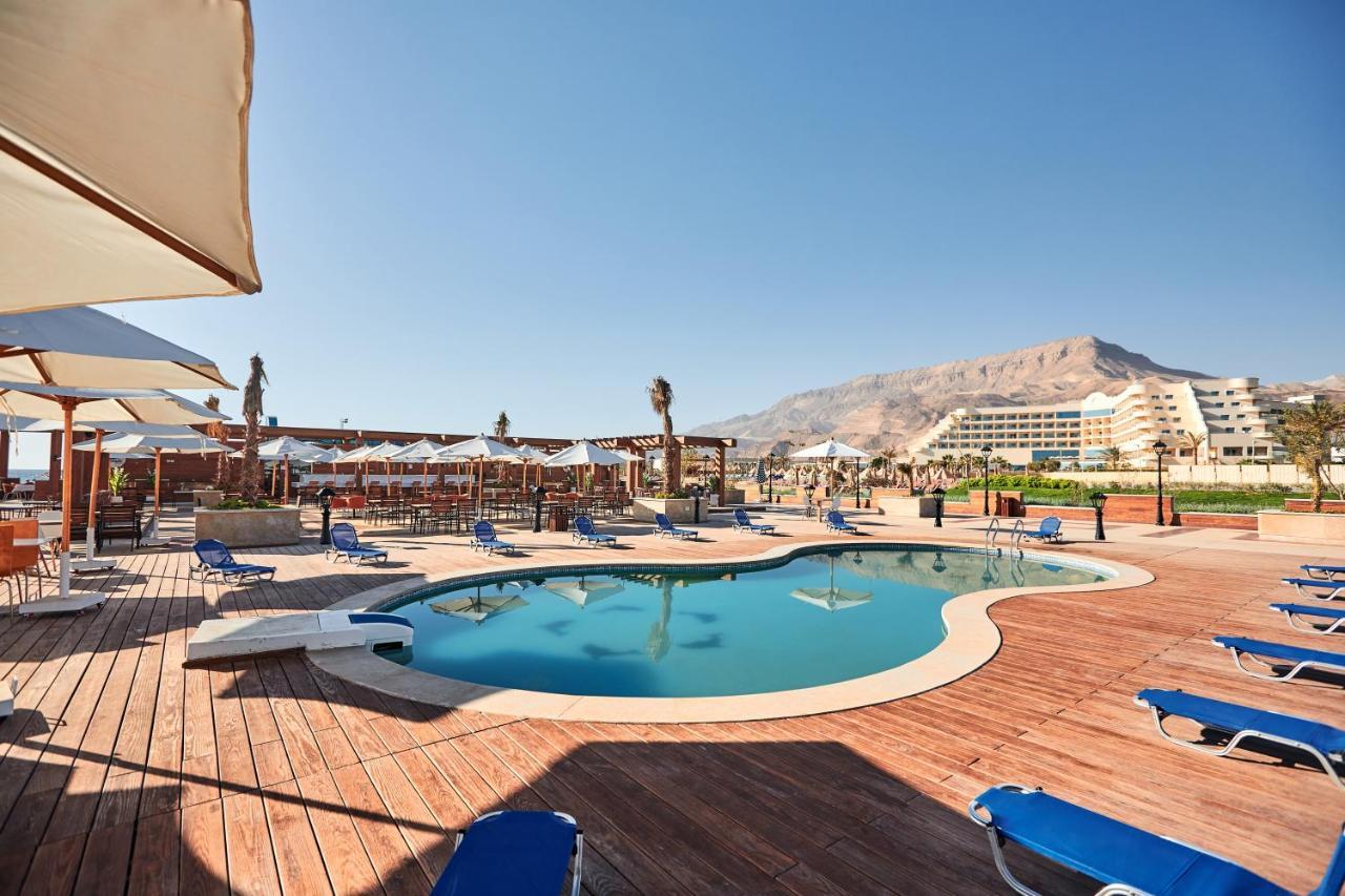 Tolip Resort El Galala Majestic Ain Sukhna Exterior foto The photo shows a swimming pool area surrounded by sun loungers and umbrellas. The pool has a unique, curvy shape and is situated on a wooden deck. In the background, there are several buildings, likely part of a resort or hotel, and a mountainous la