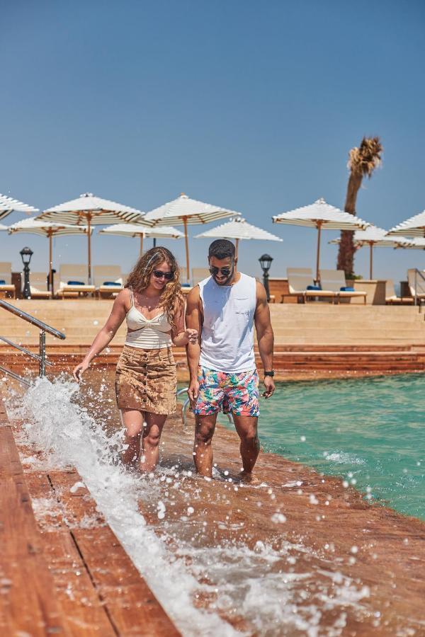 Tolip Resort El Galala Majestic Ain Sukhna Exterior foto The photo depicts a couple walking along a poolside area. They are enjoying a sunny day near a swimming pool, with splashes of water around them. The woman is wearing a light-colored top and a patterned skirt, while the man is dressed in a sleeveless