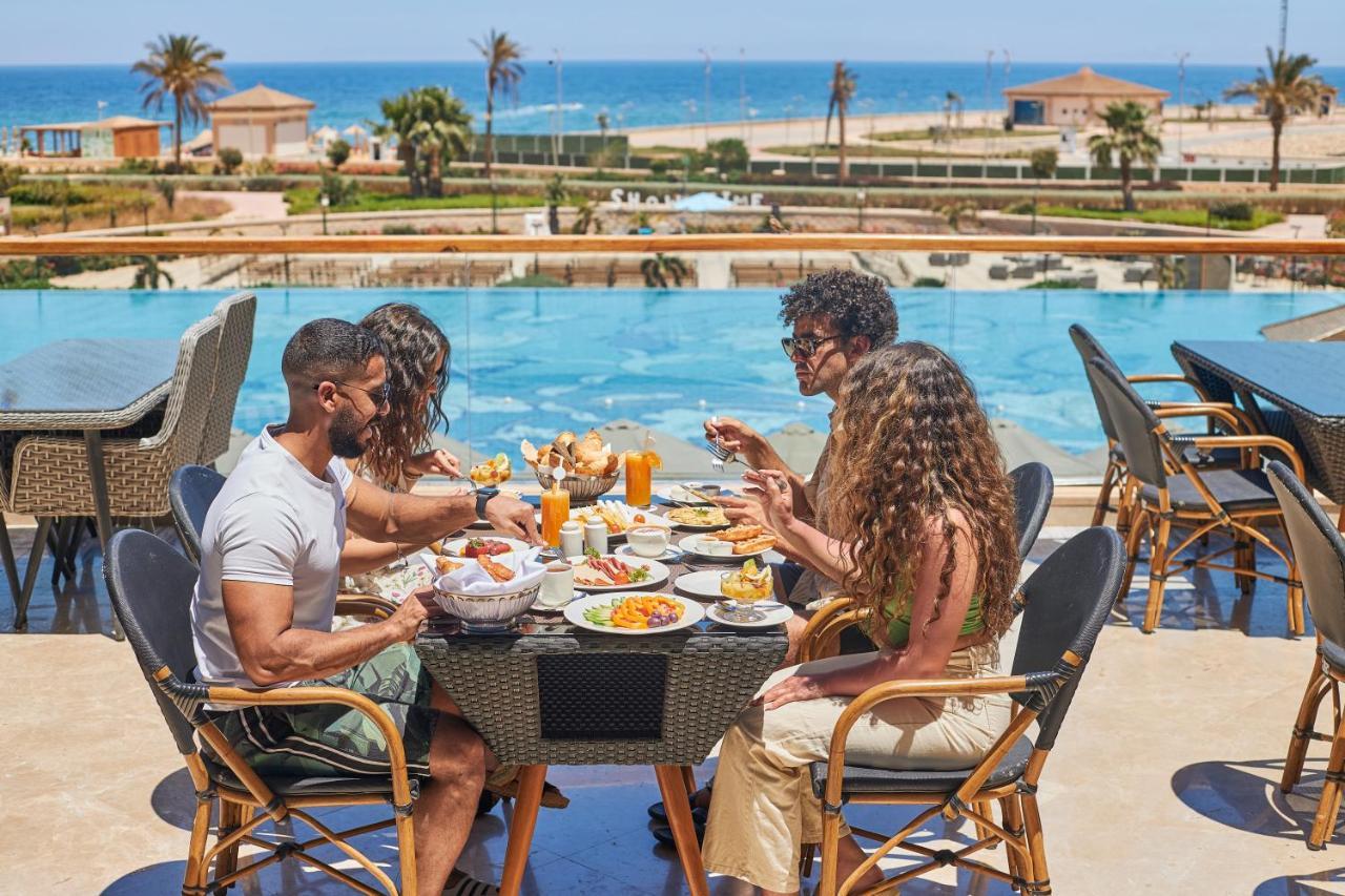 Tolip Resort El Galala Majestic Ain Sukhna Exterior foto The photo shows a group of five people enjoying a meal together outdoors, likely at a restaurant or cafe. They are seated at a table with a beautiful view of a swimming pool and the sea in the background. The table is filled with various dishes, and 