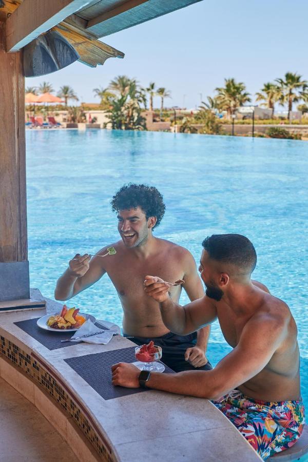 Tolip Resort El Galala Majestic Ain Sukhna Exterior foto The photo features two young men enjoying a meal together. They are sitting at a poolside table with a clear blue swimming pool in the background. The men are shirtless, suggesting a casual, relaxed atmosphere, likely in a warm environment. One man i