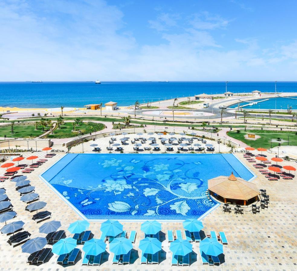 Tolip Resort El Galala Majestic Ain Sukhna Exterior foto The photo features a large swimming pool with a decorative design on its surface, surrounded by sun loungers and umbrellas. In the background, there is a view of the sea, with a clear blue sky and a few scattered clouds. The area is well-maintained, 